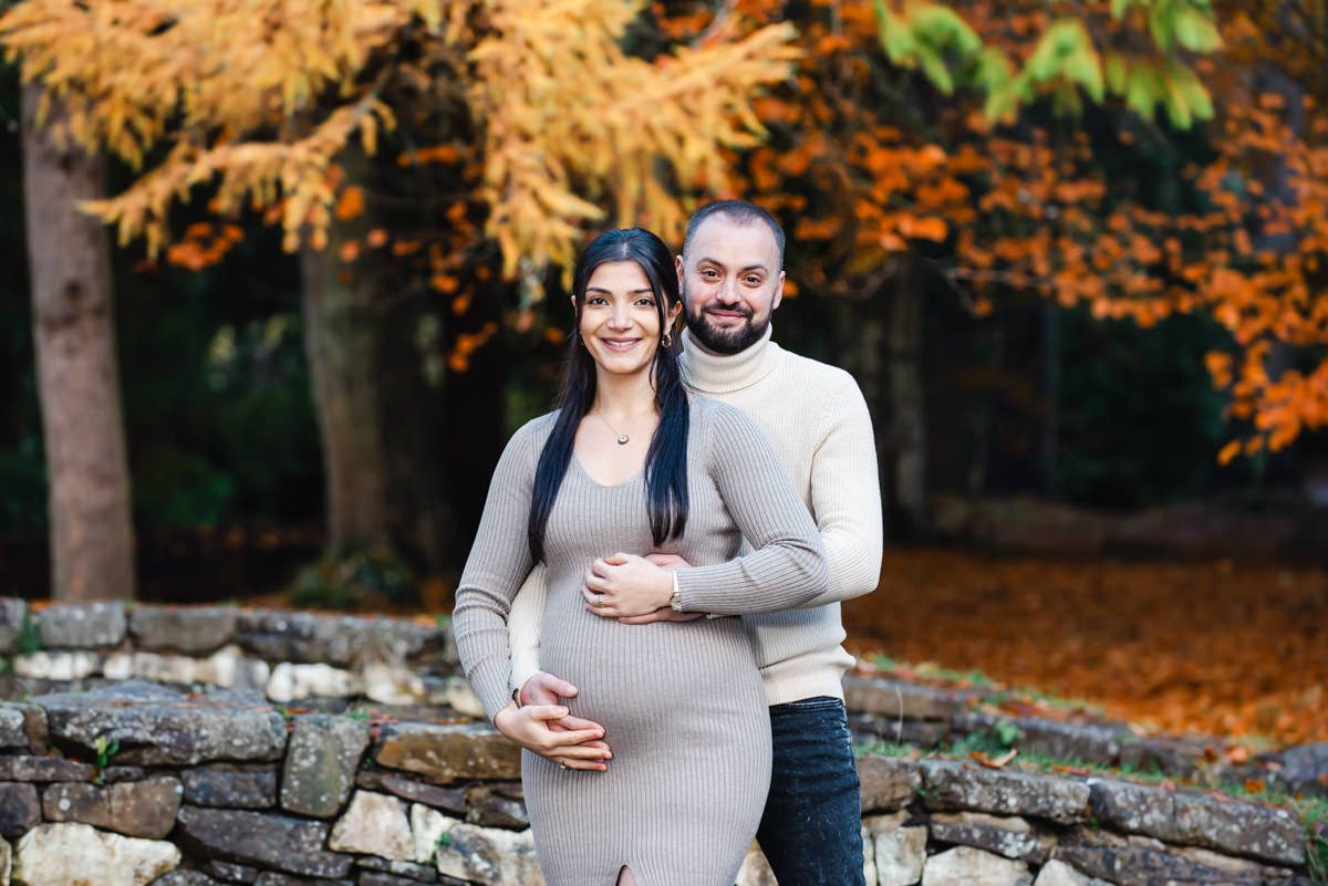 In a autumnal wooded setting a bearded man stands behind the mother to be and they both embrace the baby bump