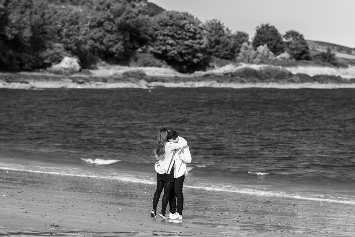 A black and white image of a young couple in pale tops and dark bottoms embracing on a beach with the sea in the background