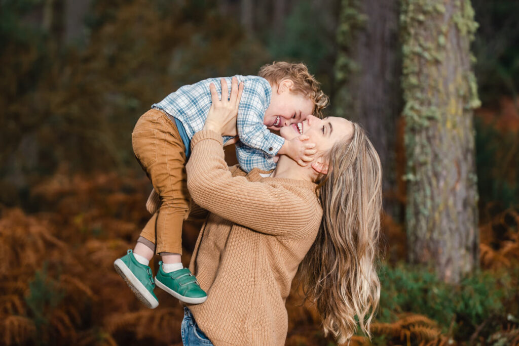 In a woodland setting a mother in a beige cardigan and blue jeans and long blonde hair holds aloft her laughing baby boy