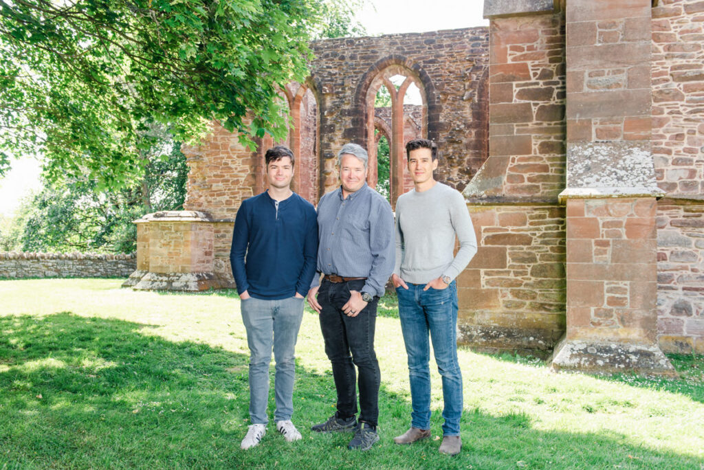 In front of a ruined stone church a grey haired father stands in between his two dark haired sons dressed in blue jeans