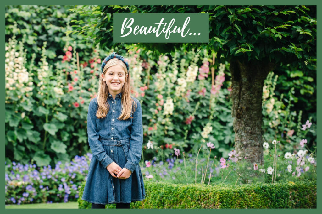 Pre-teen fair-skinned girl with long blonde hair in a denim dress and hairband smiling in a garden with flowers and a tree