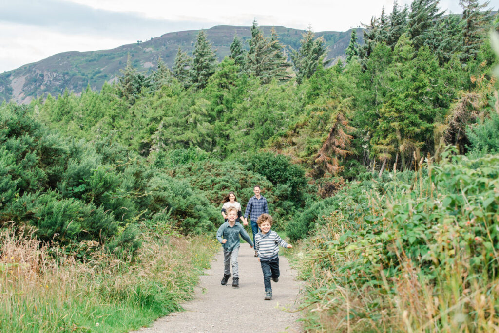 In a coniferous woodland setting two young boys run along a dirt path ahead of their parents in the background