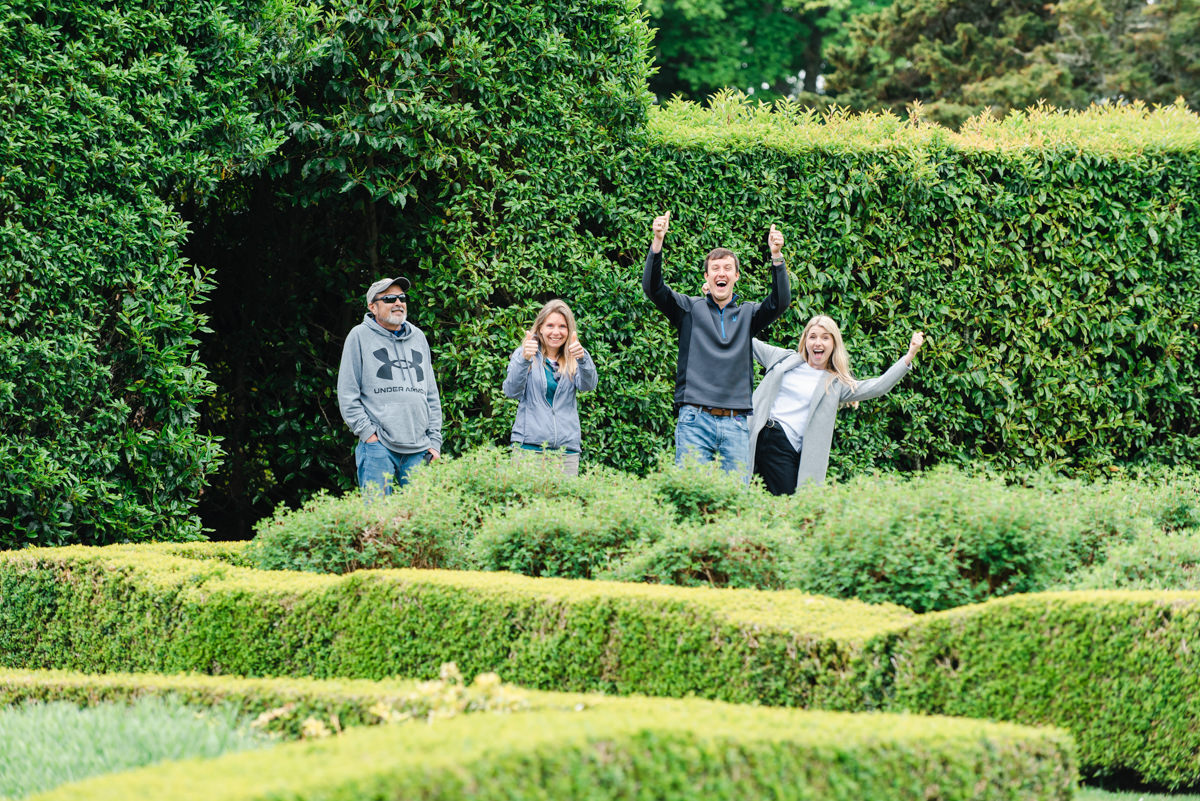 A group of people cheer with raised hands while standing in a mature evergreen garden with large hedges