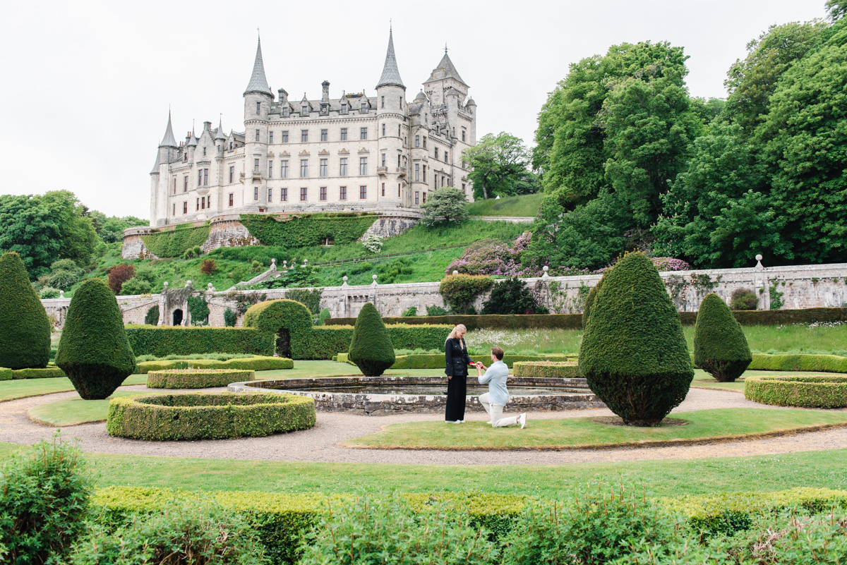 In front of a large castle in a mature garden setting while on one knee a man in light clothing proposes to his fiance