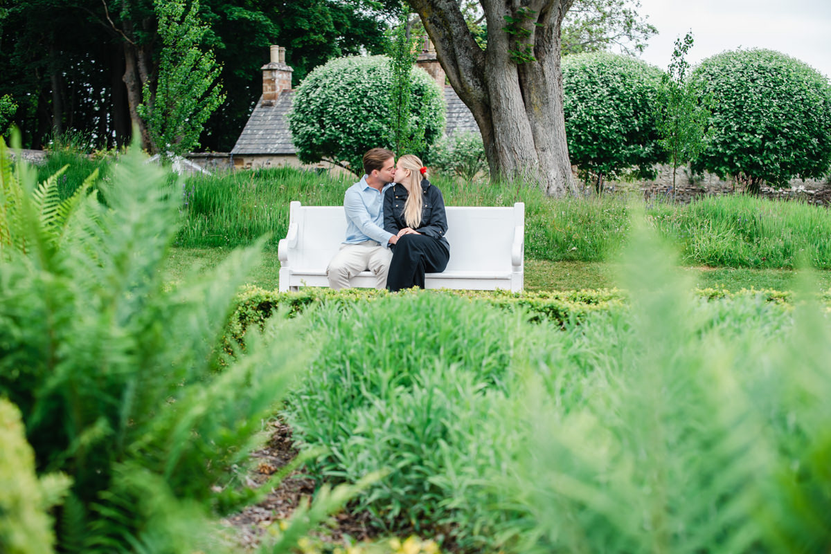 A young couple sit on a white bench and share a kiss in the middle of a mature evergreen garden