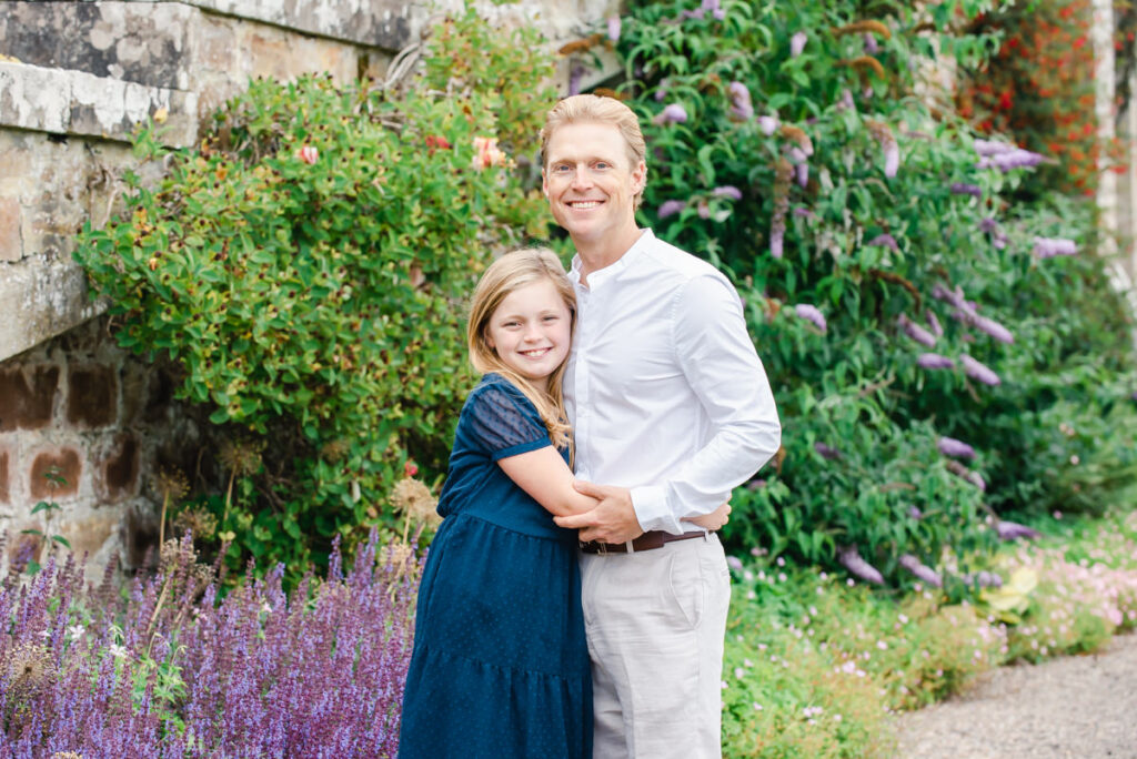 A father dressed in white hugging his pre-teen daughter in a blue dress in a garden with a stone wall and purple flowers