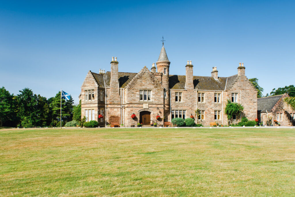 A side view of a large sandstone manor house sitting in front for a large green lawn below a clear blue sky