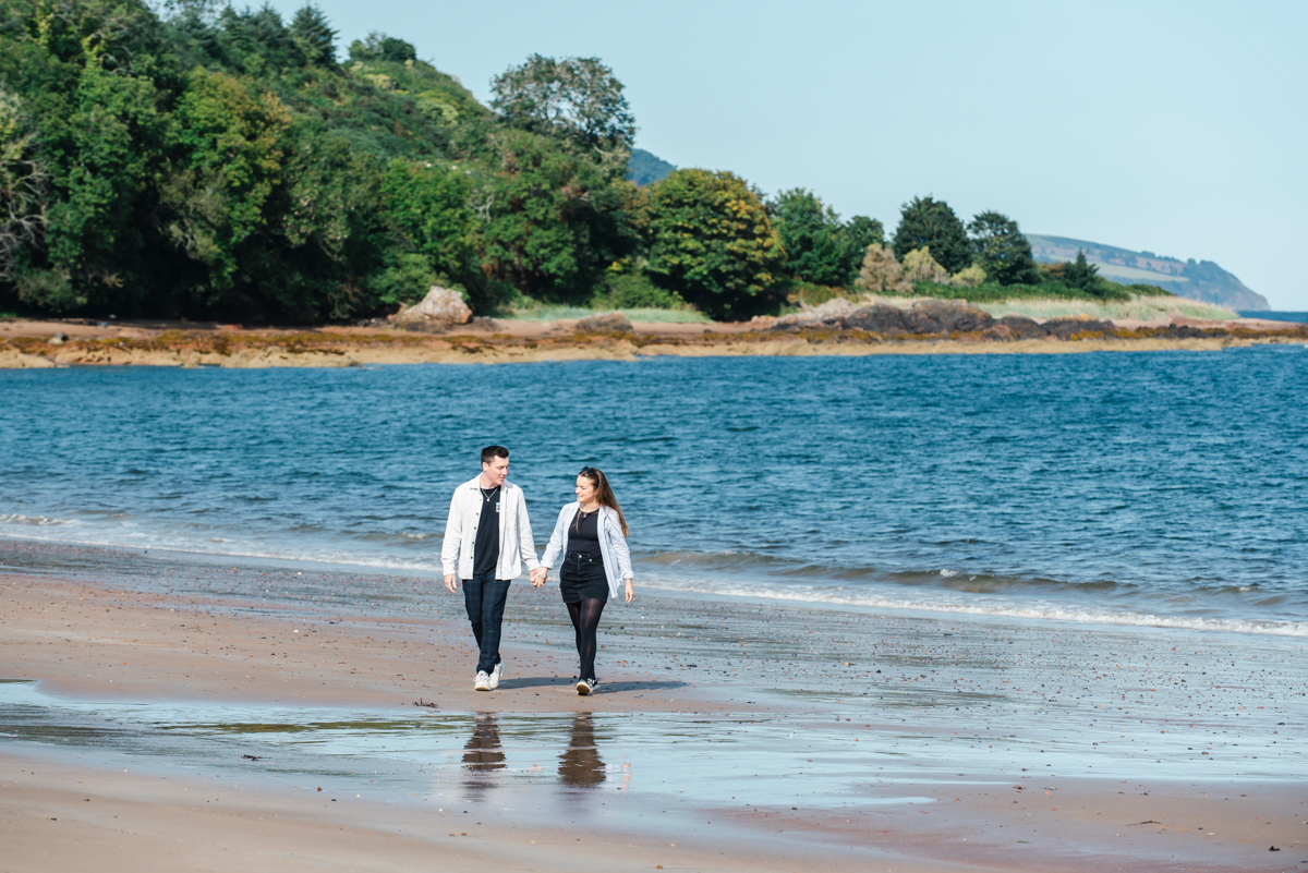 A young couple in pale tops and dark bottoms chat and hold hands while walking along a beach with the sea in the background