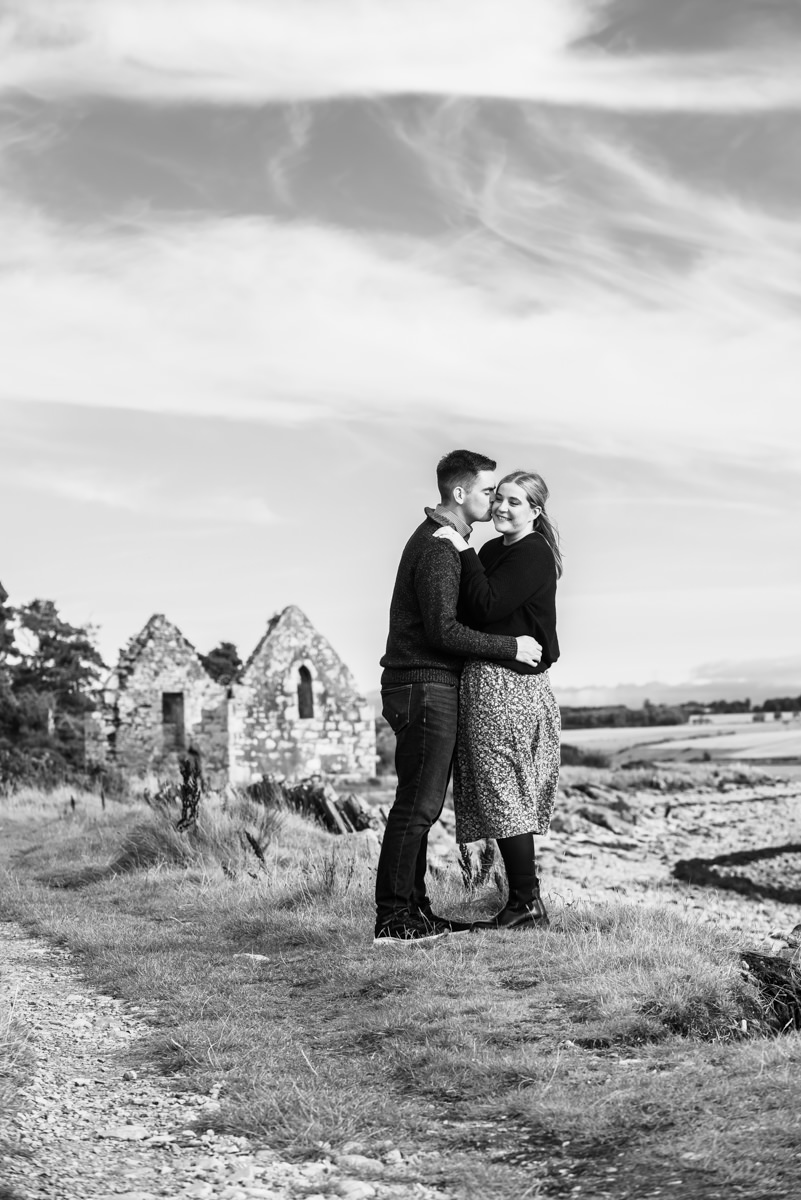 A black and white image of a man below a dramatic sky kissing a woman on the cheek in front of a ruined chapel by the shore