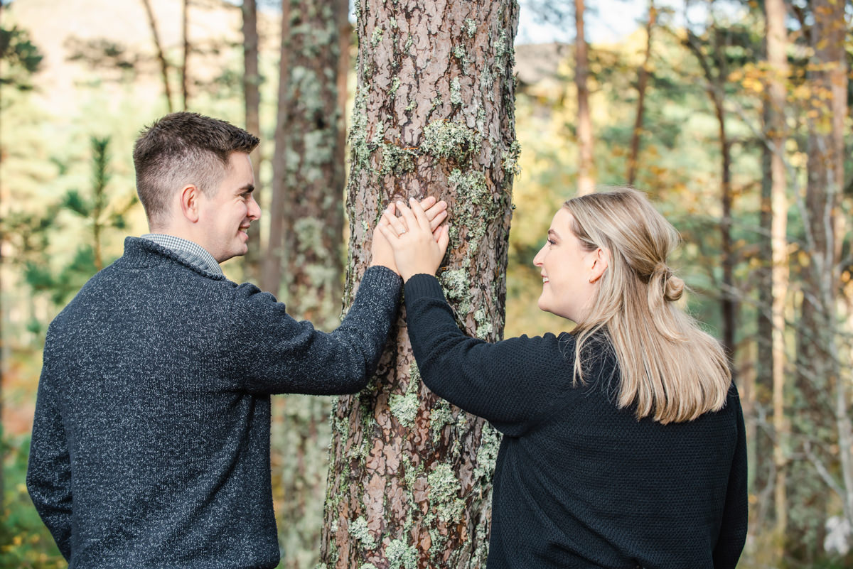 In a coniferous woodland setting a young couple put their hands on a tee to show off their new engagement ring