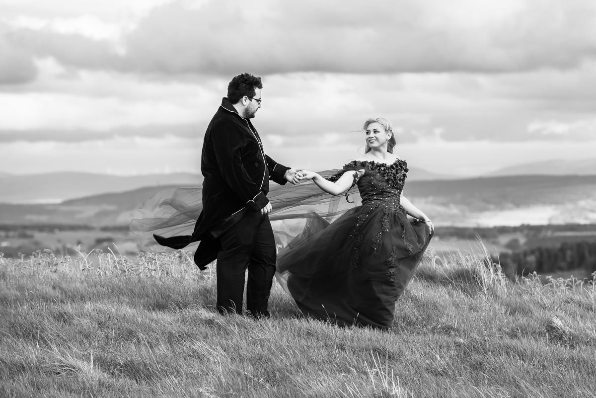 A woman in a long flowing dress with a cake holding hands and looking romantically at a man in a suit on a hilltop