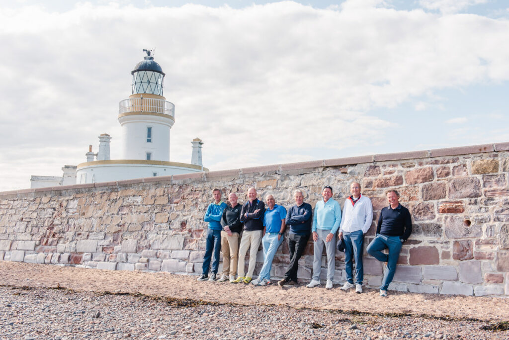 A group of eight mature men lean against a large sandstone wall in front of a white lighthouse on a stoney beach