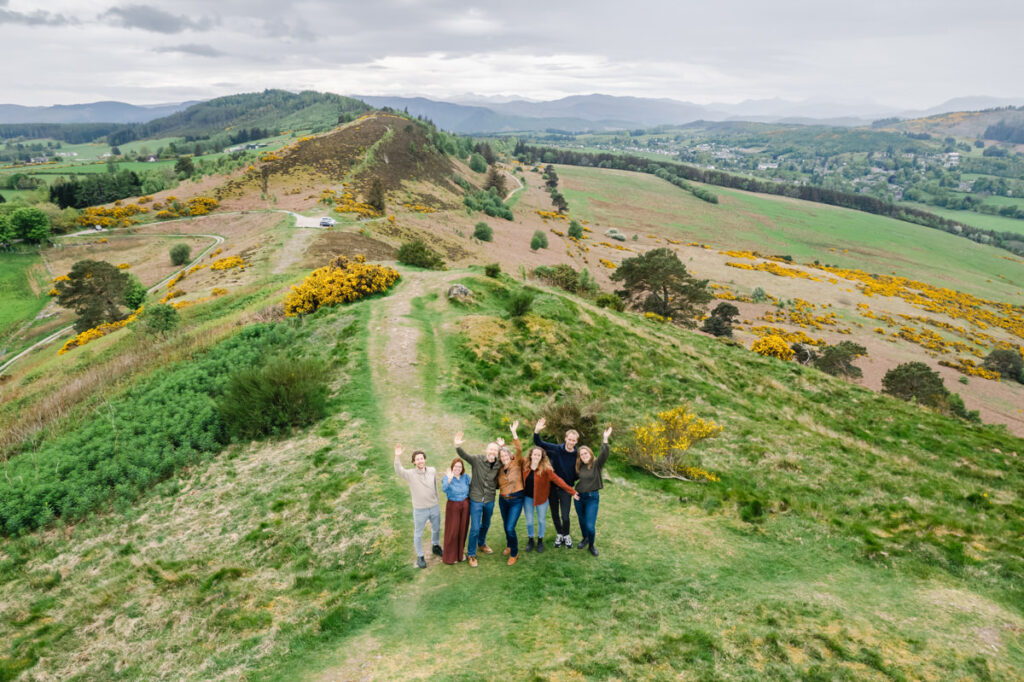 An elevated shot of a family of seven looking up and waving from a small hill in a highland setting in spring
