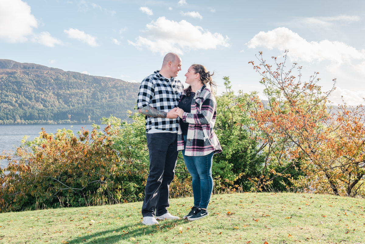 On a grassy knoll overlooking a loch and hills a young couple in checkered tops hold hands and smile at each other