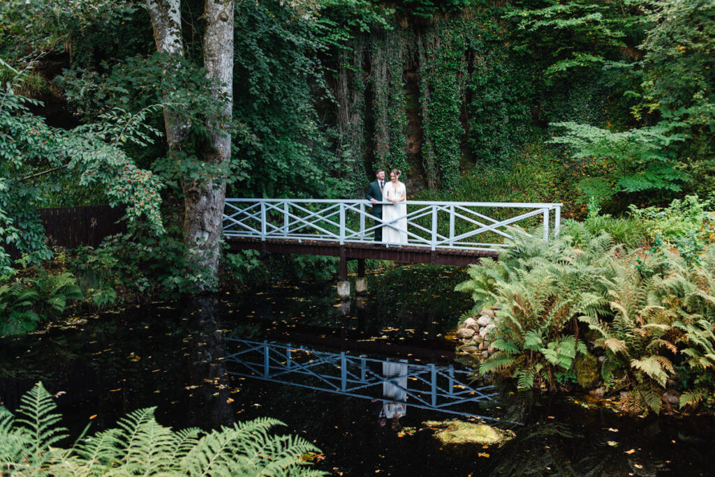 In a woodland setting a bride and groom stand on a white bridge that spans across a dark pond that shows their reflection