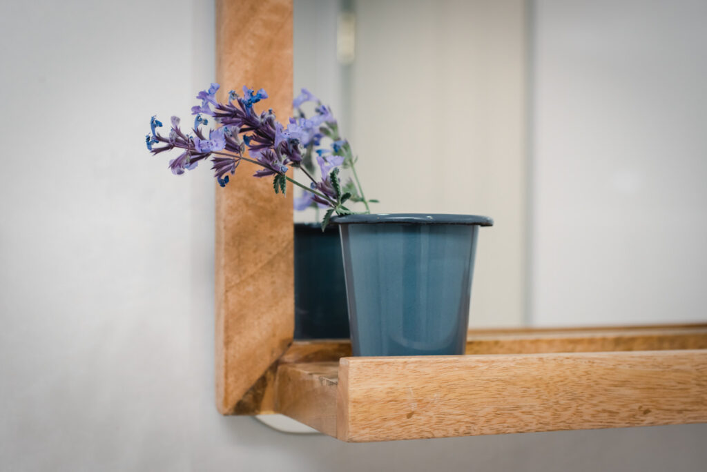 A close up of lavender in a grey pot sitting on a wooden shelf underneath a wooden framed mirror