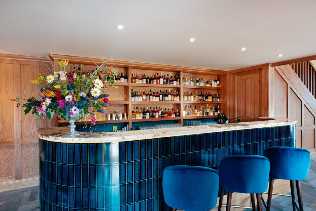 A blue gloss tiled bar island with blue stools and wooden shelving containing many bottles of malt whiskey