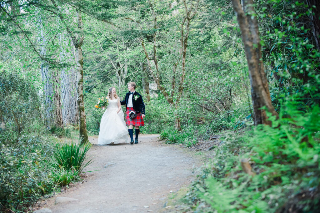 A groom in a red kilt and dark jacket walks with his bride who is holding a floral bouquet along a woodland path