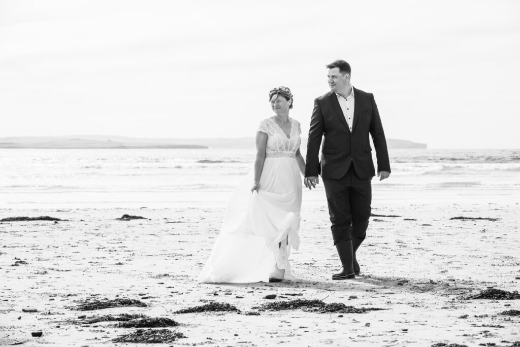 A monochrome image of a bride and groom walking hand in hand along a beach on a sunny but windy day