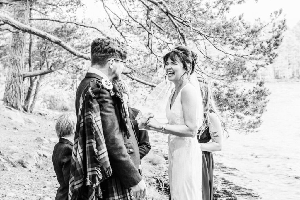 A monochrome image of a married couple in a woodland setting by a lochside sharing a funny moment