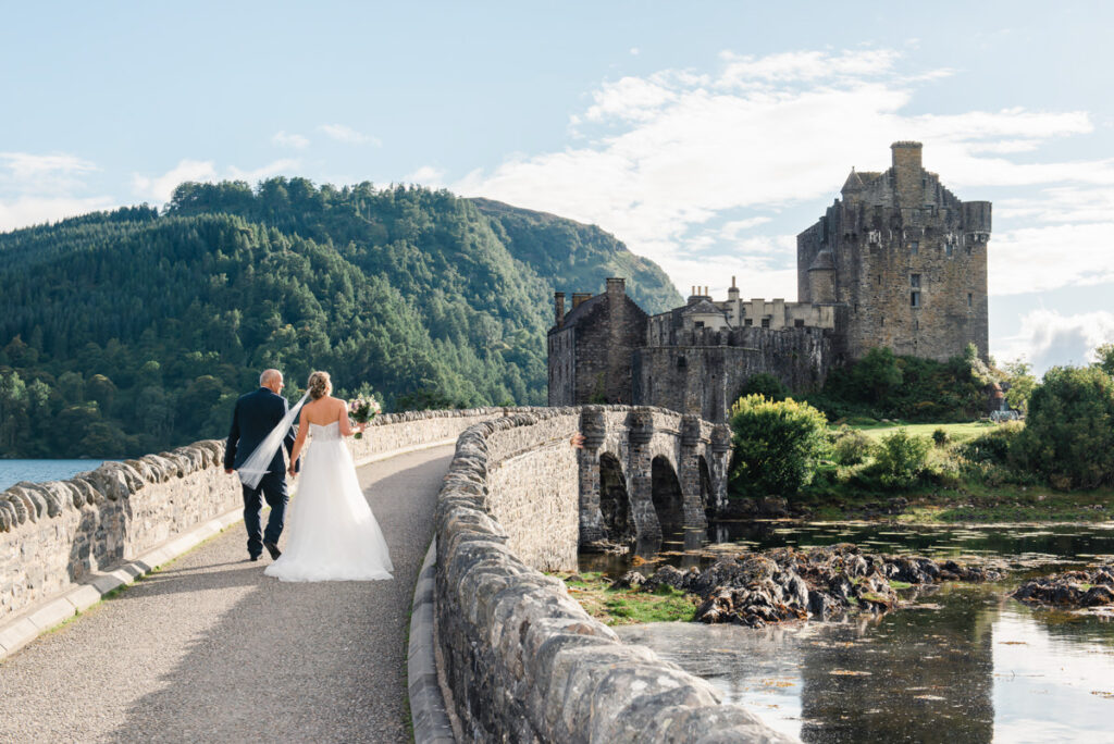 A bride and groom walk along an old stone bridge that leads to a large Scottish castle by a loch