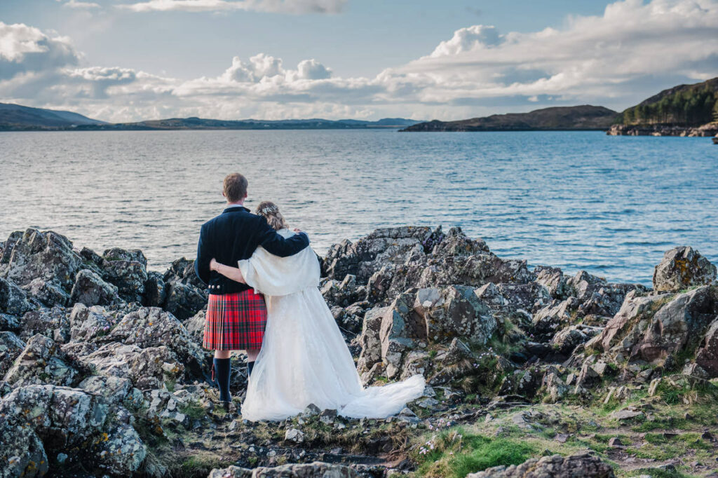 A man in a red kilt and a woman in a wedding dress embrace while looking out toward an island in a coastal setting