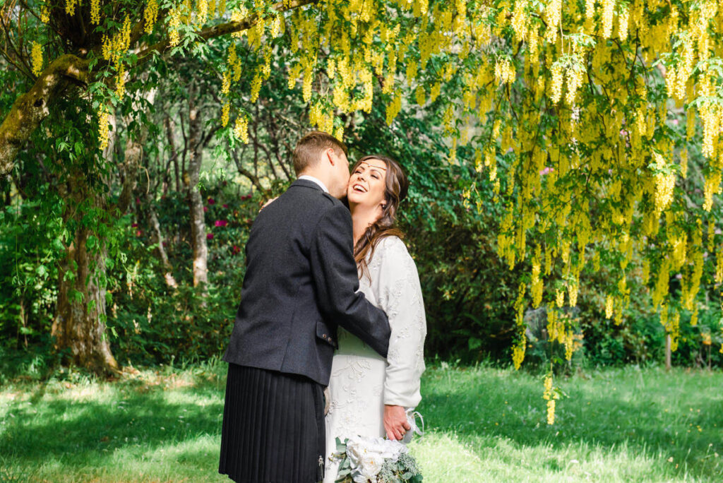 Under a yellow laburnum tree a groom dressed in a blue kilt kisses his long brown haired bride on her cheek