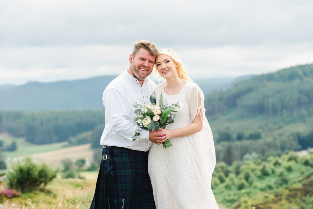 In a highland setting a man with a blonde beard,hair and blue kilt embraces his wife dressed in a white wedding dress and shawl