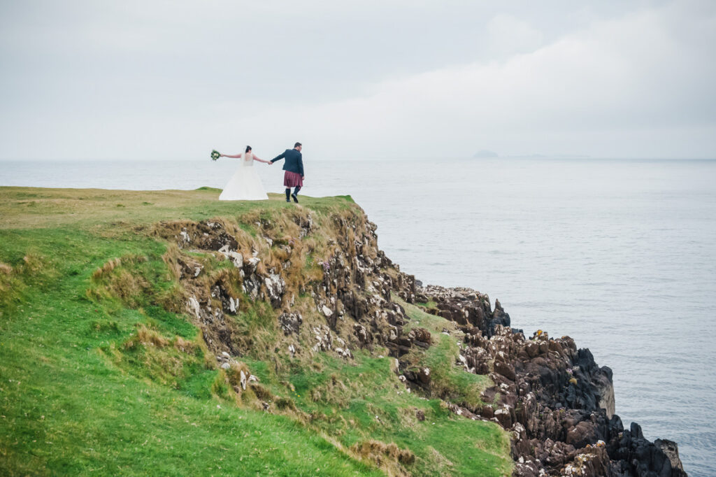 On the edge of a grassed cliff a married couple walk hand in hand looking out toward the sea under a grey sky