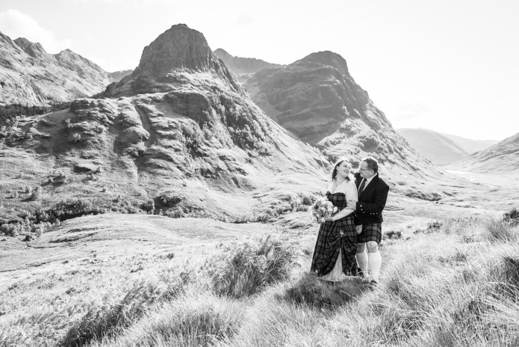 A monochrome of a newly wedded couple embracing on a high-point overlooking a Scottish mountain range