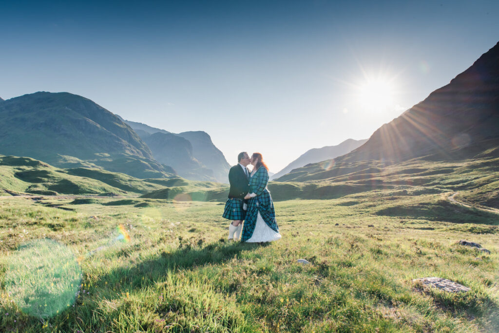 A bride and groom in traditional tartan dress share a case surrounded by Scottish mountains