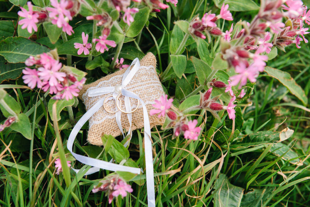 A close up of a pillow shaped jute wedding favour wrapped in white ribbon sitting on grass next to a red campion plant