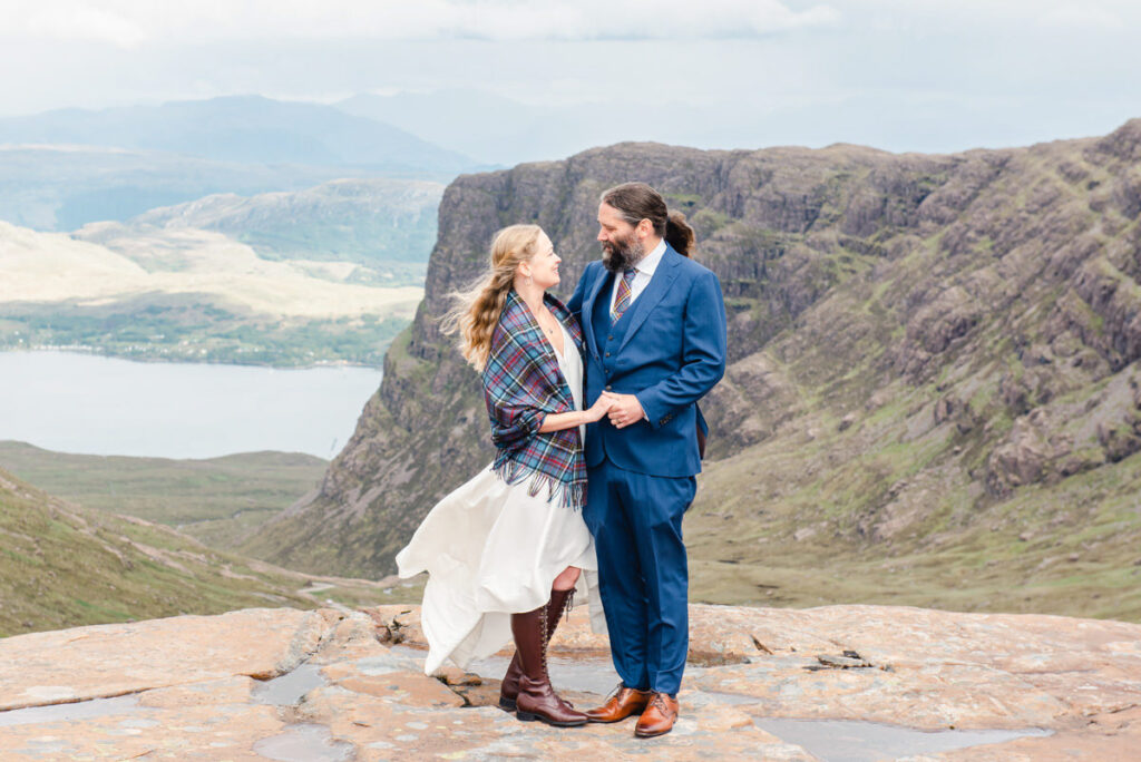 A bearded groom holds the hands of his bride wearing a blue tartan shawl while standing on a cliff over looking a mountains