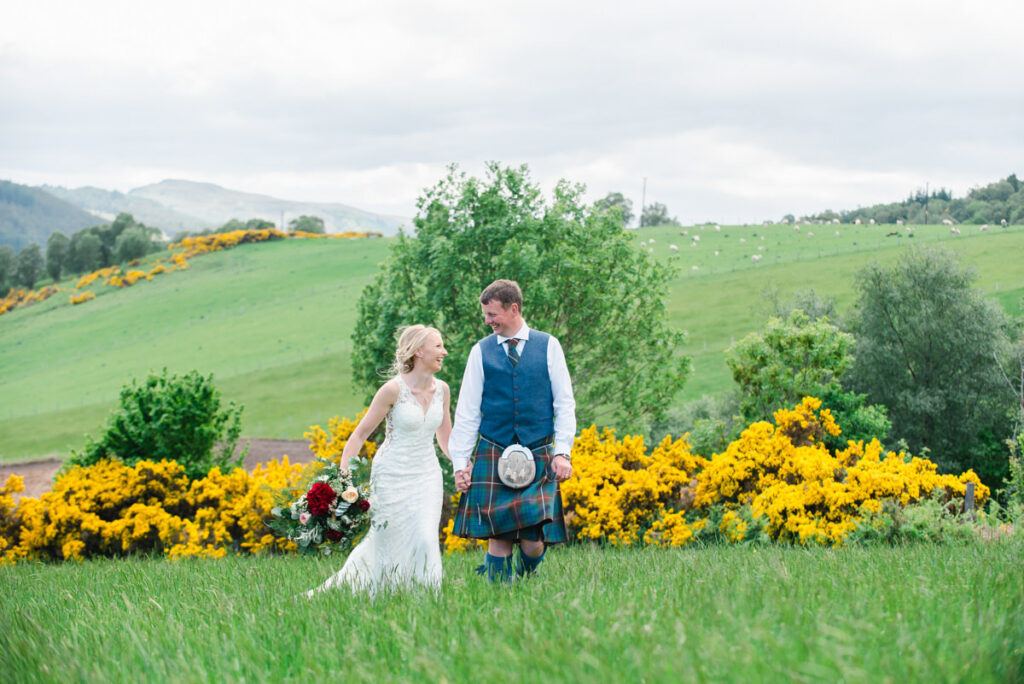 A bride and groom share a laugh while walking through a grass field in front of a large golden gorse bush