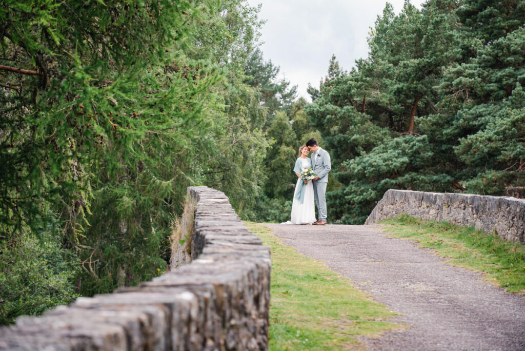 A newly wedded couple stand and embrace in the middle of a stone bridge with coniferous woodland all around
