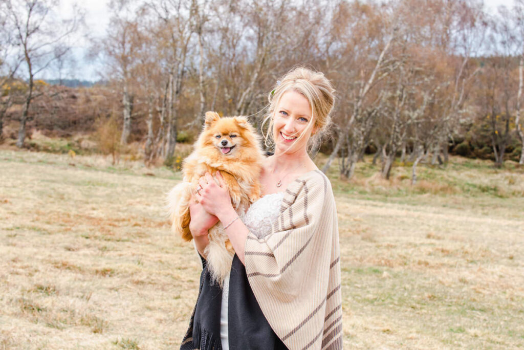 In a autumnal outdoor setting a bride in a blue dress and beige shawl holds up her small dog to the camera and smiles