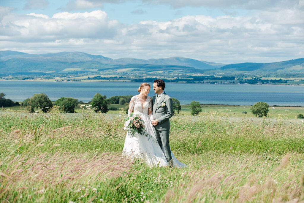 A bride and groom embrace in the middle of a large grassy field overlooking water and a blue sky