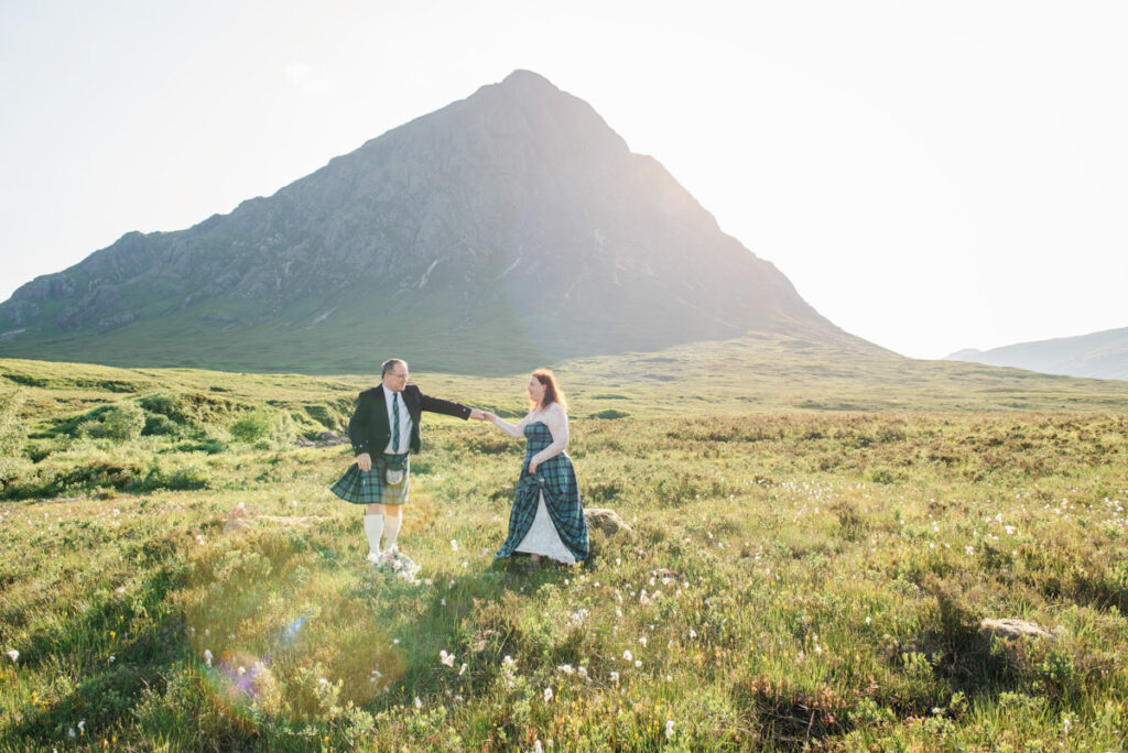 A bride and groom share a dance in the middle of a heather heath in front of a large mountain in the sunshine