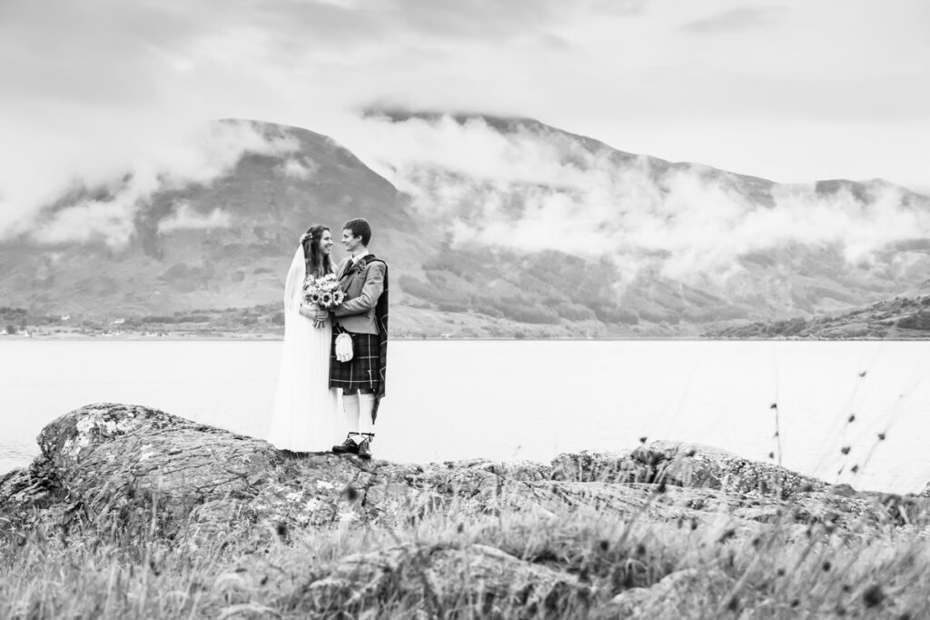 A monochrome image of a bride and groom standing on a stony outcrop before a loch and misty mountain top