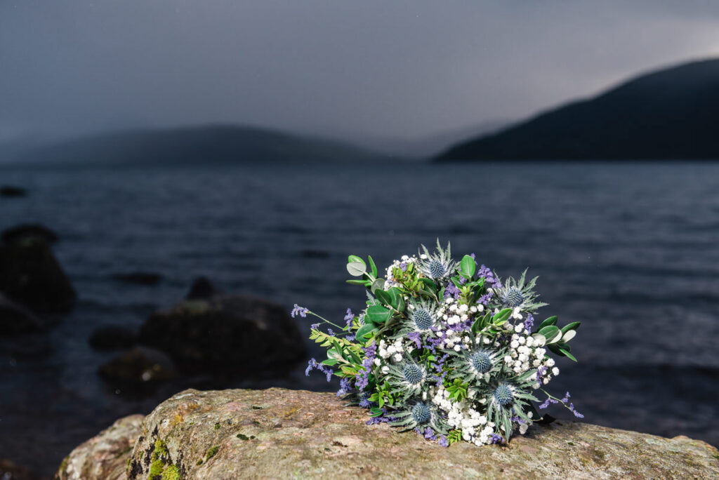 A close up of a blue thistle and white heather wedding bouquet sitting on a rock in front of a dark and moody loch