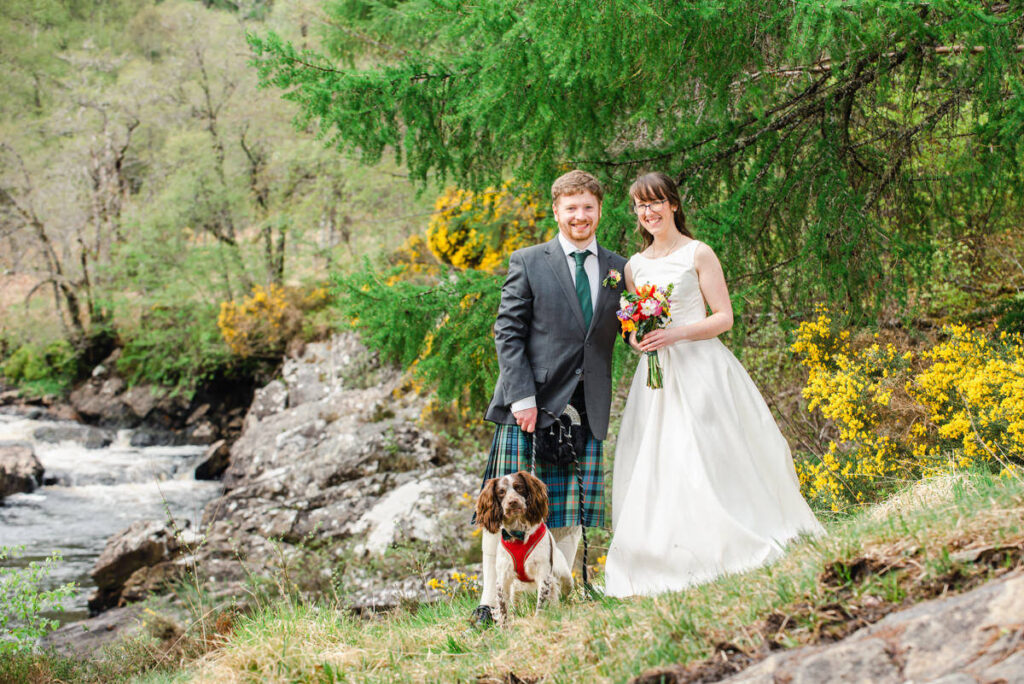 A man with a red beard, green kilt and tie, stands next to his wife in a white wedding dress with their dog at their feet