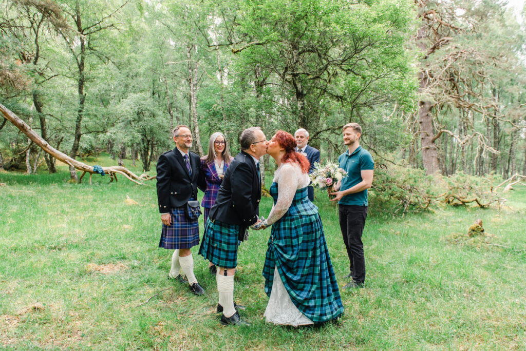 In a woodland setting a bride and groom dressed in green tartan share a kiss while the wedding party look on
