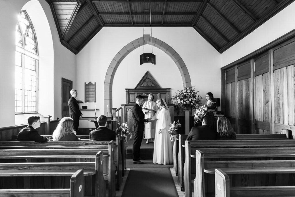 A monochrome image of a bride and groom standing at the alter of a small chapel exchanging rings while the family look on