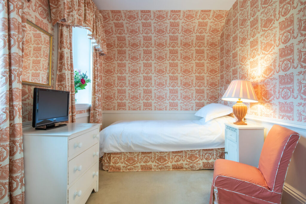 A single white bed sits at the back of a room with peach coloured printed wallpaper and white furniture