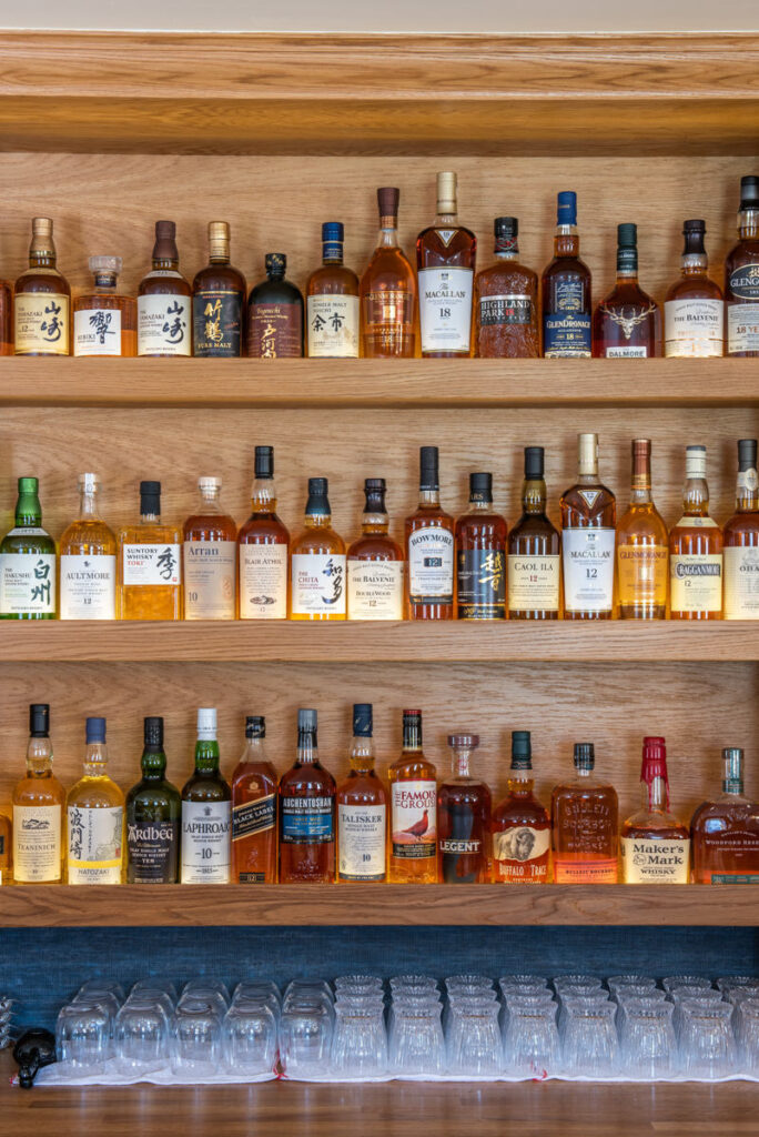 A close up image of many many bottles of whiskey neatly stacked on wooden shelves above clear whiskey glasses