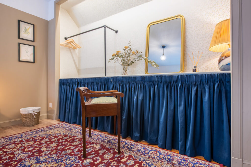 A large gold mirror sits on a dresser with a blue curtain at its base in a room with white walls and a wooden chair