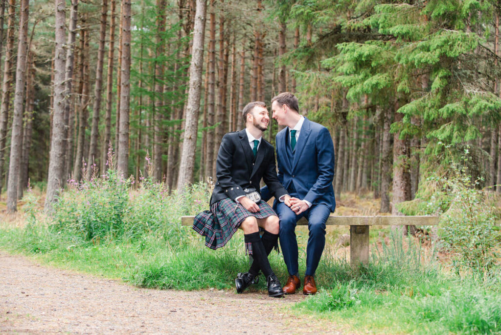 In a woodland setting a tall man in a blue suit looks into his husbands eyes and shares a tender moment while holding hands