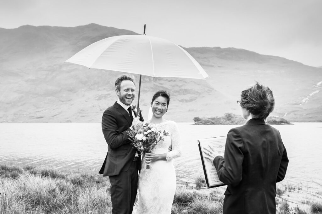 A monochrome image of a married couple under a white umbrella being read their vows while the wind blows the celebrants hair