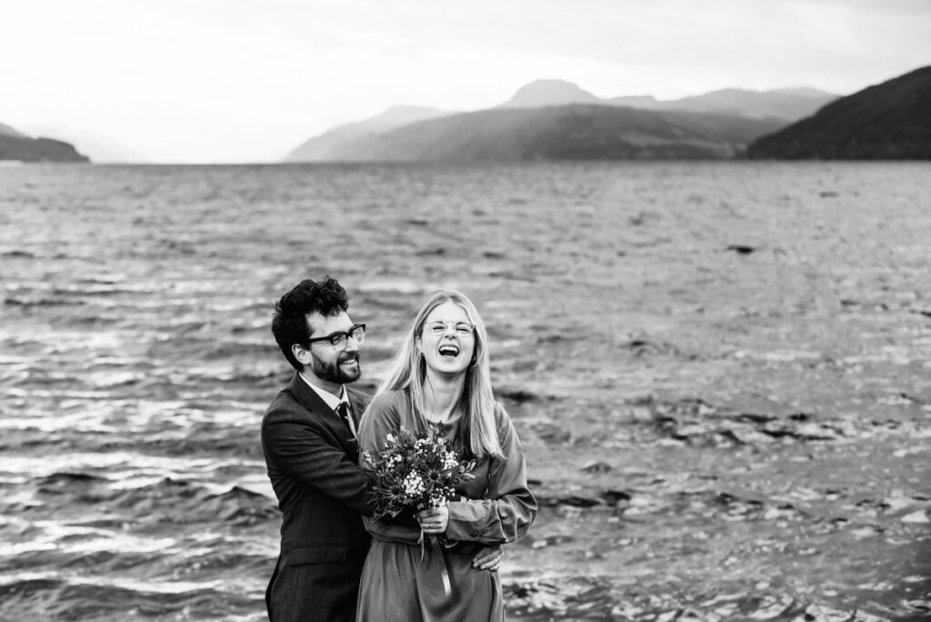 A monochrome image set by a loch of a bearded groom in glasses embracing his bride who is holdings her bouquet and laughing