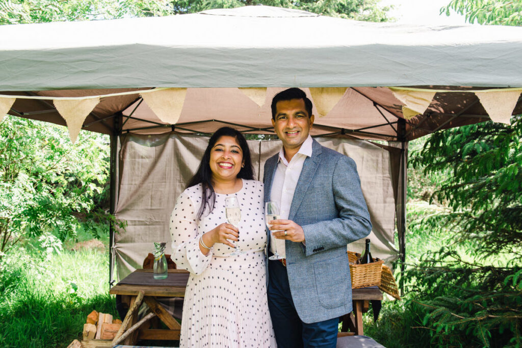 In a woodland setting a dark skinned couple stand under a gazebo while toasting their marriage with a glass of champagne